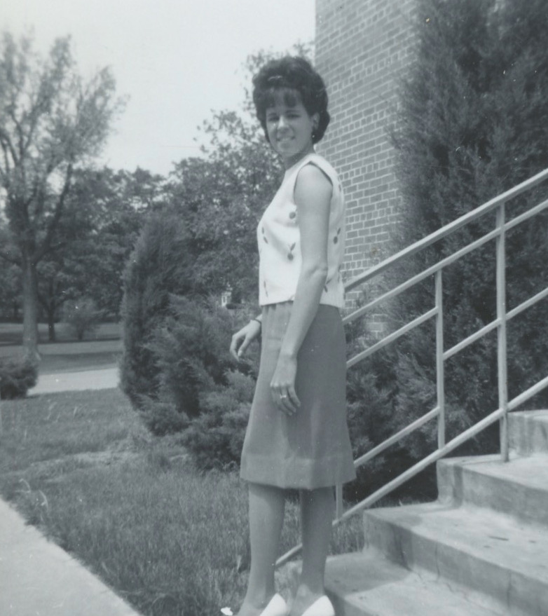 A young white woman with wavy dark hair stands on some outside stairs. Her arm is on the railing and her head is turned toward the camera as she steps down. She's wearing a light-colored sleeveless top and a slim skirt.