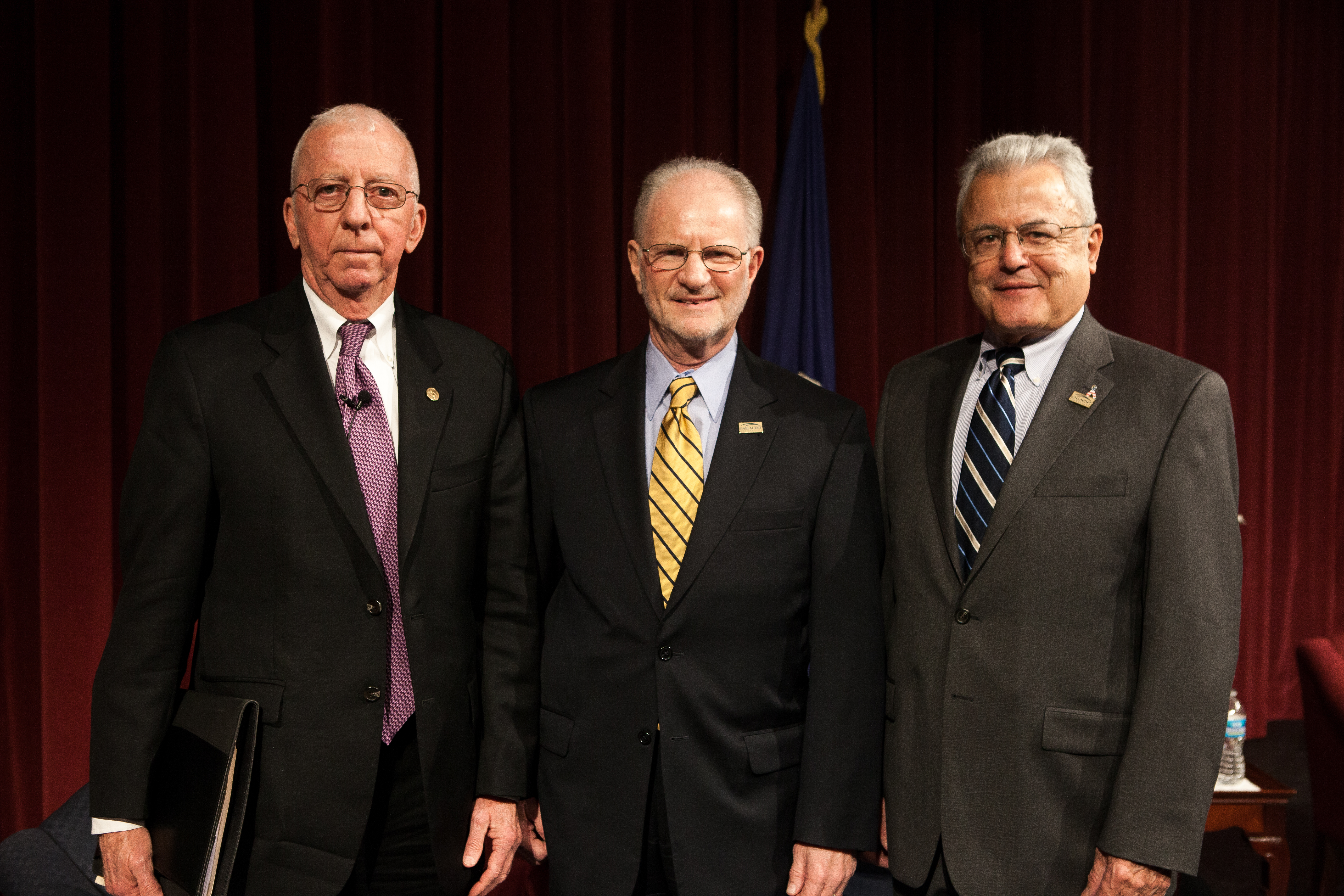 Three older men in dark suits. Two are white and one is Latino. All wear glasses and label pins. 