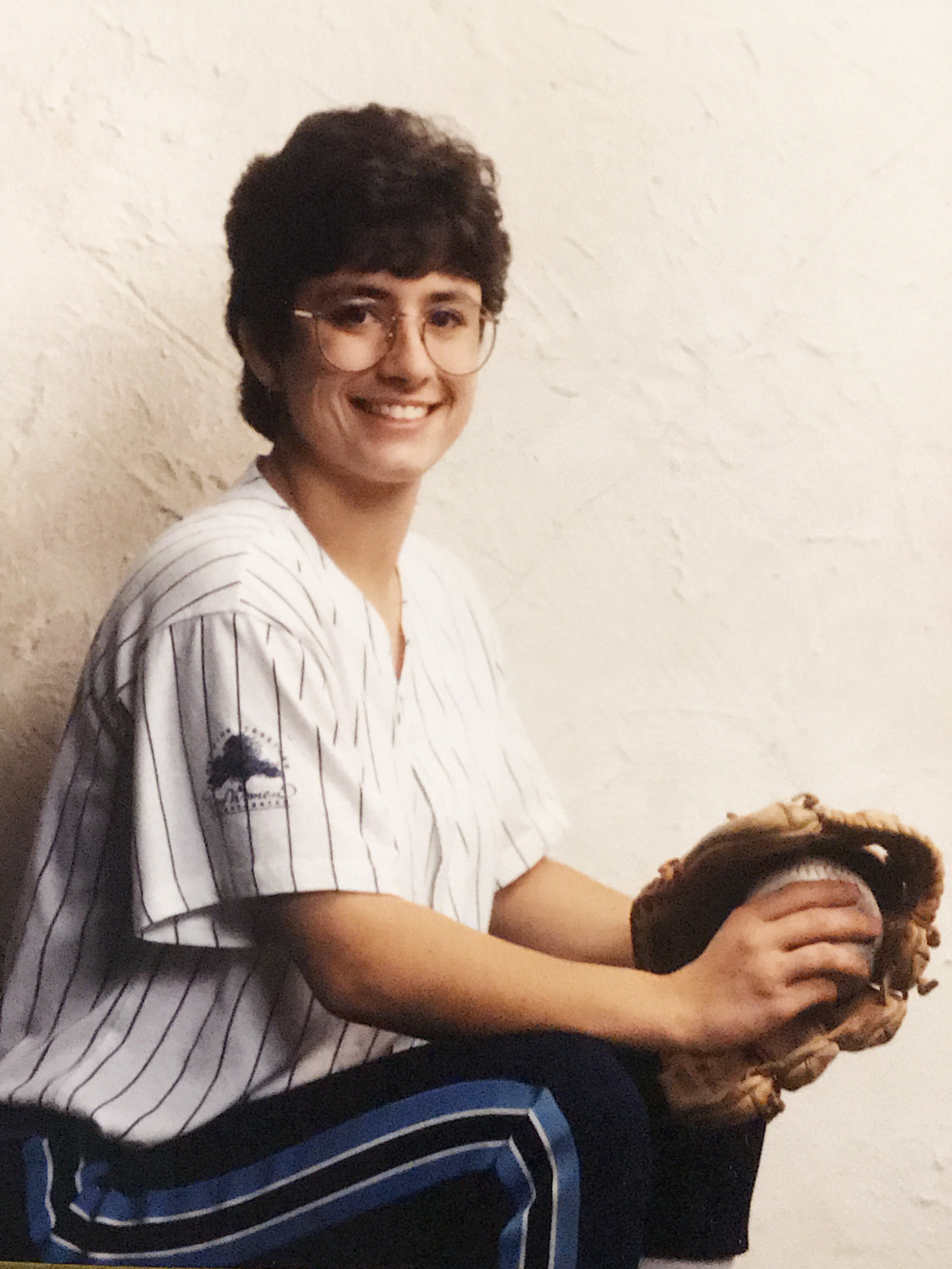 A young white woman with wavy dark brown hair, large round glasses, wearing a softball uniform holds a ball in a glove.