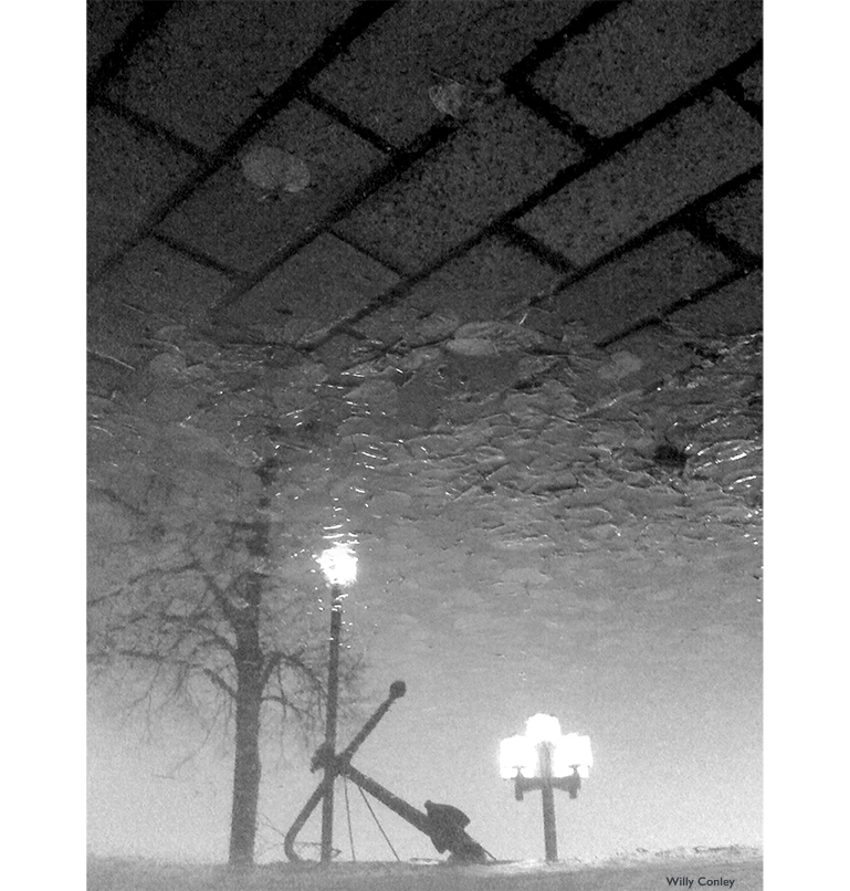 black-and-white vertical photo of a partial water reflection over a brick-paved surface. The main features being reflected in the water are a large ship anchor, a couple of lit streetlamps, and a bare tree. Seen through the reflection at the bottom of the puddle are fallen leaves.