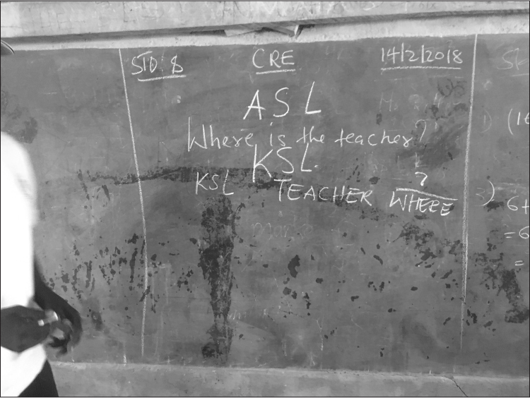 Chalk writing on a marked and stained blackboard; a black person in a white T-shirt is standing half off-camera, holding chalk. The writing on the board reads “STO 8, CRE, 14/2/2018” across the top, and then “ASL” and “Where is the teacher?” in the center. Below is written “KSL” and “KSL TEACHER WHERE”.