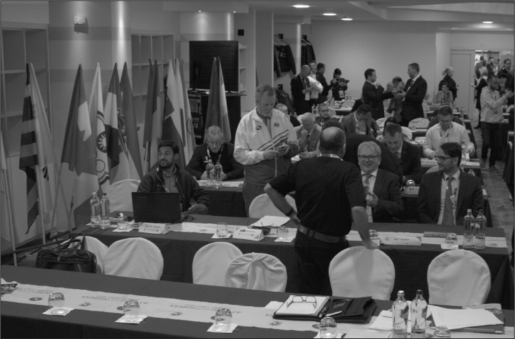 Interior of a room with rows of long trestle tables with green tablecloths and delegate packs, with flags lining the side. People in suits or sportswear are sitting and standing around socializing.