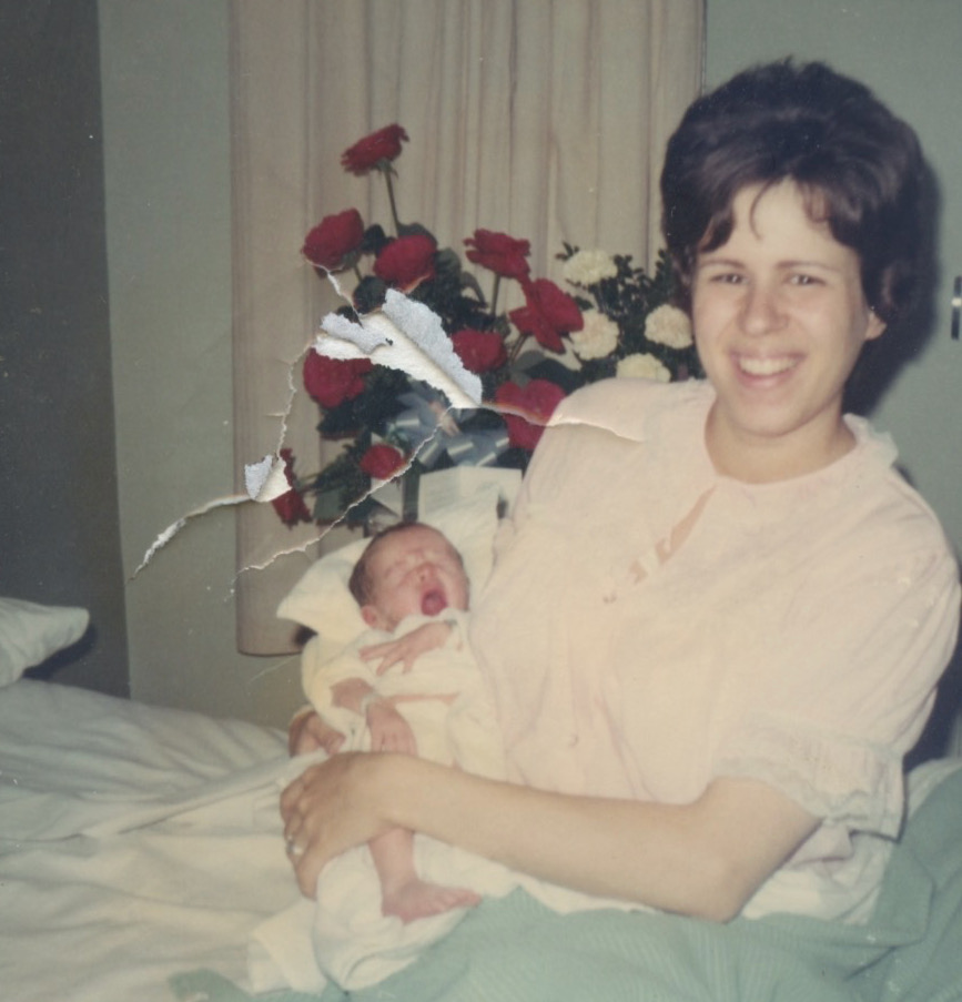 A smiling young white woman in a pink dressing gown sitting up in a hospital bed holds a white newborn baby in her arms. Behind her is a large bouquet of red and white roses.Image
