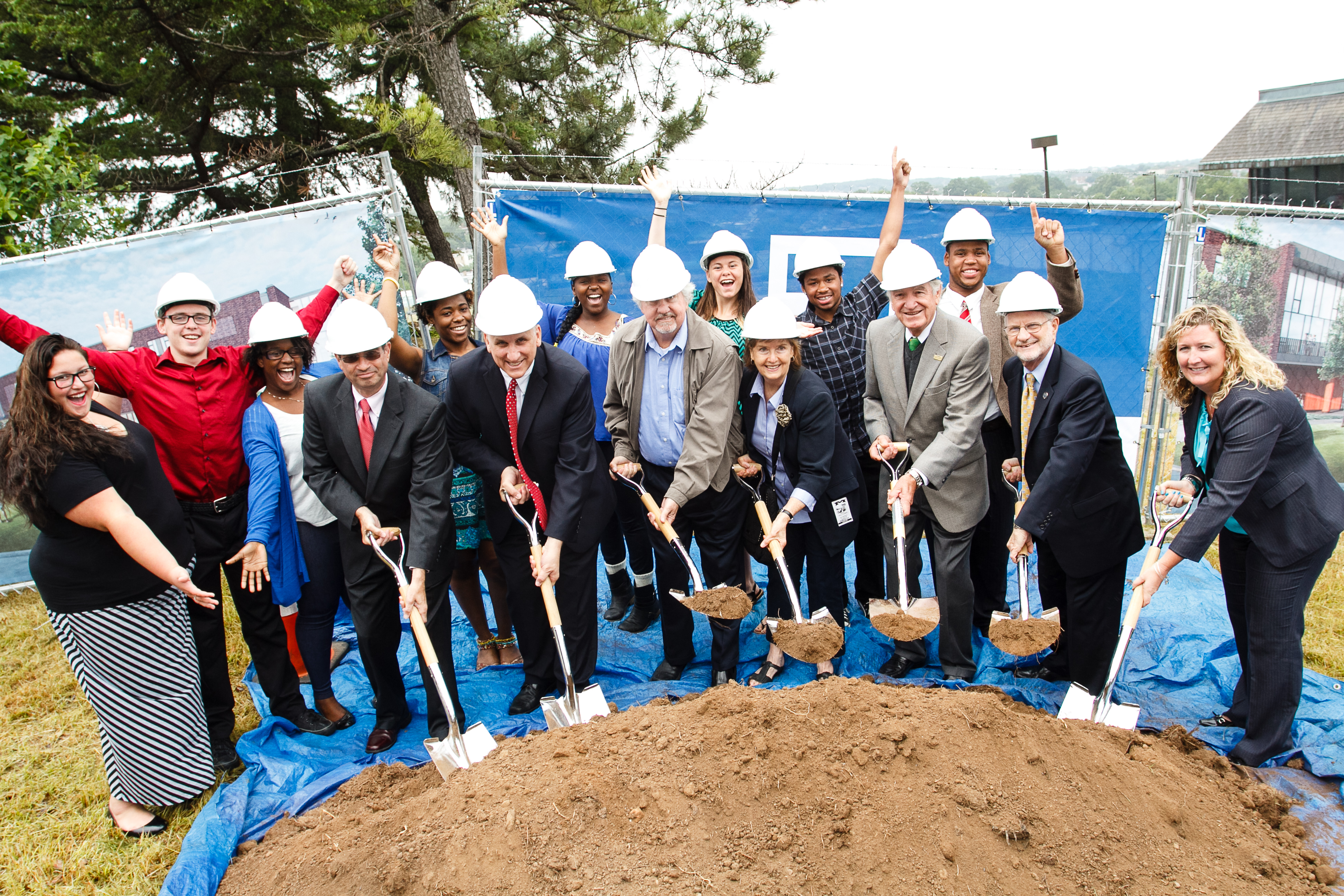 A group of white adults wearing white hard hats and holding shovels in front of a large pile of dirt. Some are digging into the pile while others already have dirt in their shovels. Behind them a group of younger people, five Black students and three white,  some also wearing hard hats, cheer the adults on.