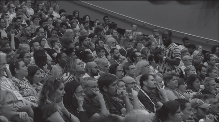 A filled conference hall. Everyone is seated except a man in the foreground, who is standing up in his seat, facing away from the stage and signing.