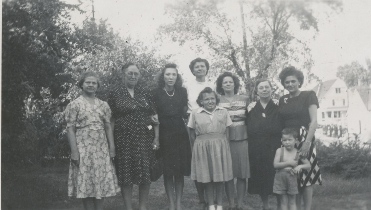 A black and white photo of a group of eight white womn of various ages standing in front of a screen of trees. the young woman on the far right has her hand on a young boy in a jumper. The women all wear dresses.