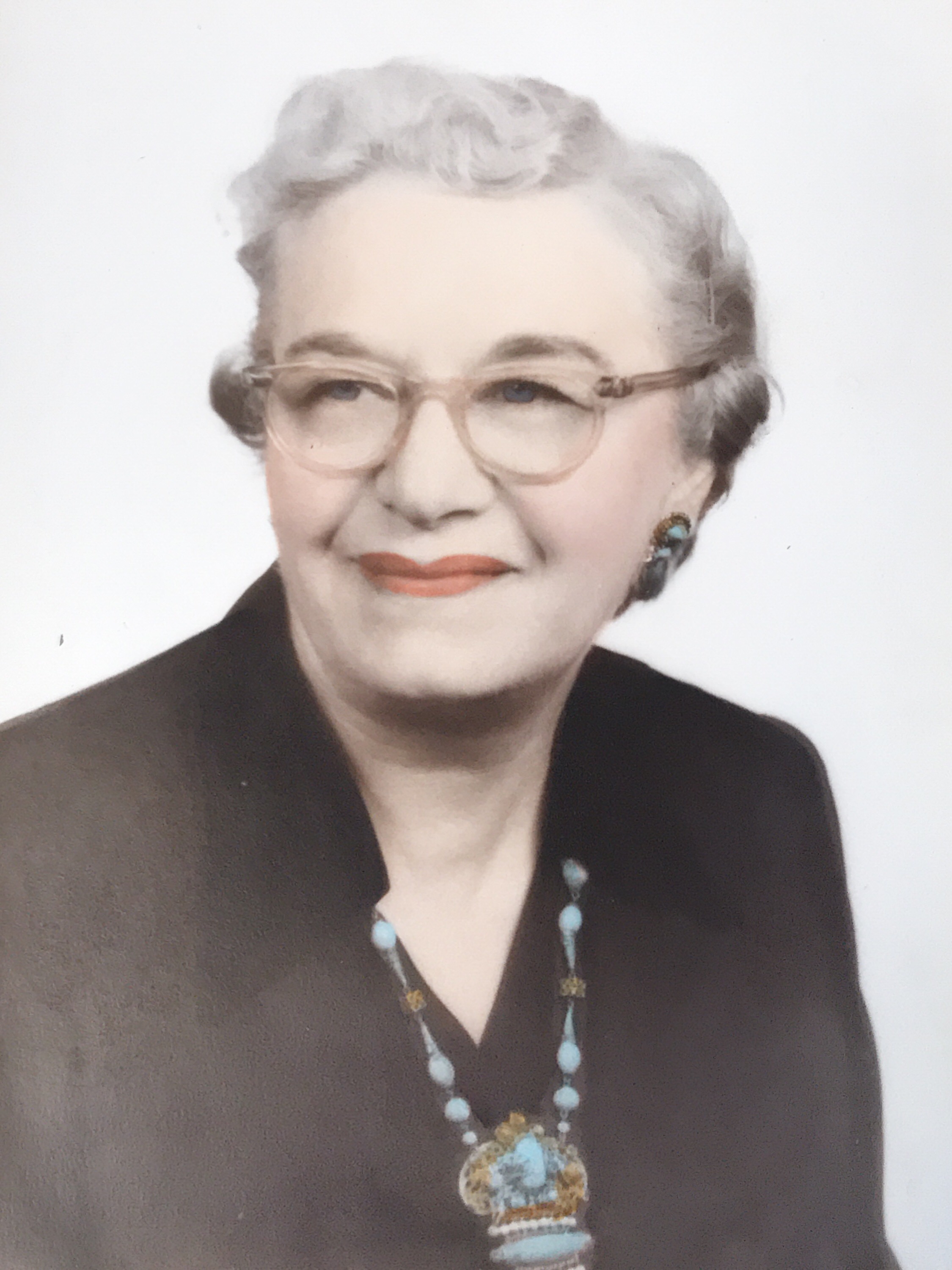 A color photo of a white-haired older white woman with glasses, looking to the left. She's wearing light blue-and-black earrings and a beaded necklace.