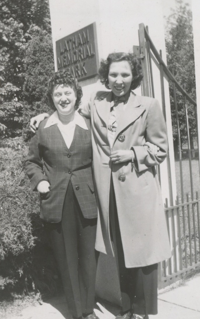 Two young white women in front of a gate that has the words Latham Memorial Park attached to the pillar it's connected to. The women are both smiling at the camera. The shorter woman, on the left, is wearing a pants suit and has one hand in her jacket pocket. The other woman has her arm around the shorter woman and is wearing a long light-colored coat. Both women have wavy dark-colored hair.