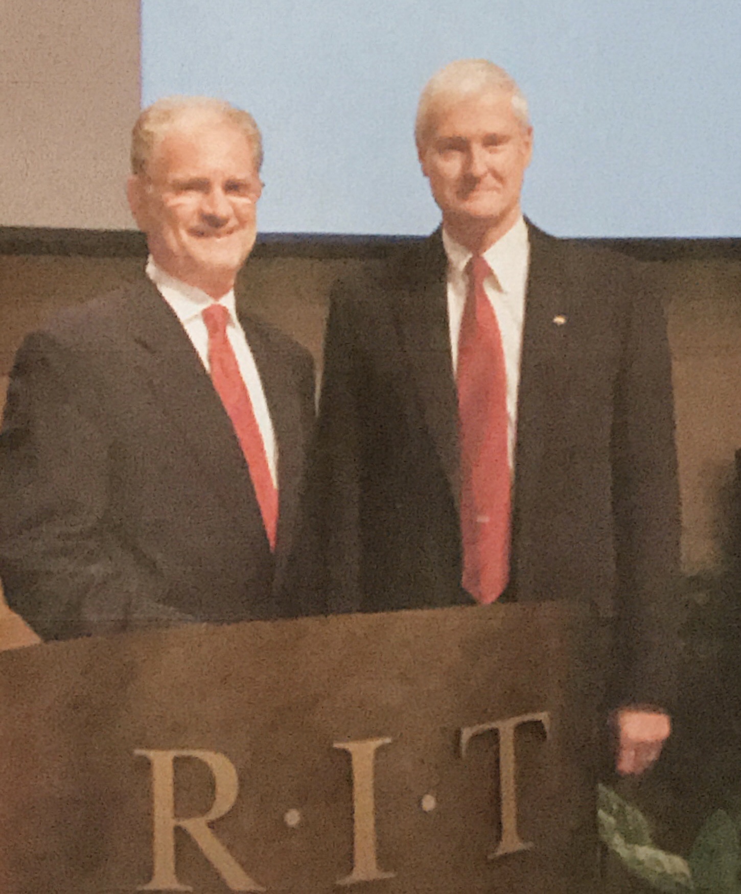 Two middle-aged white men both in dark suits with red ties stand behind a podium with the letters R-I-T across the front. The man on the left is shorter and is wearing glasses. The man on the righ is slender and tall with white hair.