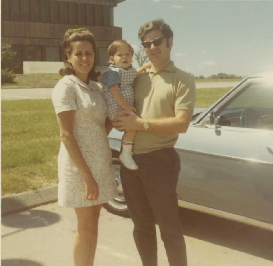 A young white woman with thick brown hair pulled back and in a short light-colored dress stands next to a young white man wearing sunglasses, a polo shirt and long pants. He's holding a little white boy dressed in a blue checkered outfit between them. Behind all of them is a powder blue car.