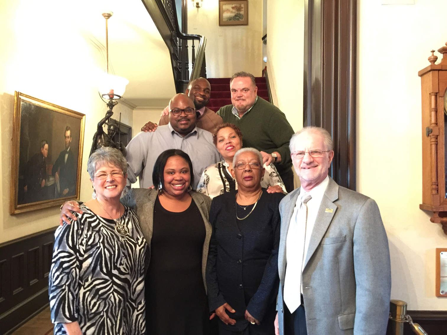 A group of eight people standing in front of and on a staircase. Across from the stair case is a painting of Abraham Lincoln. The people standing are an older white woman in a white and black top, a young Black woman with an olive jacket over a black top, an older Black woman with white hair and glasses, an older white man in a light gray suit and light colored tie. Behind them is a young Black woman in a cream and black top, a middle-aged Black man in a gray shirt. Directly behind him, with a hand on his shoulder, is a young Black man in a brown top. Next to him is a middle-aged white man wearing an olive v-neck sweater.
