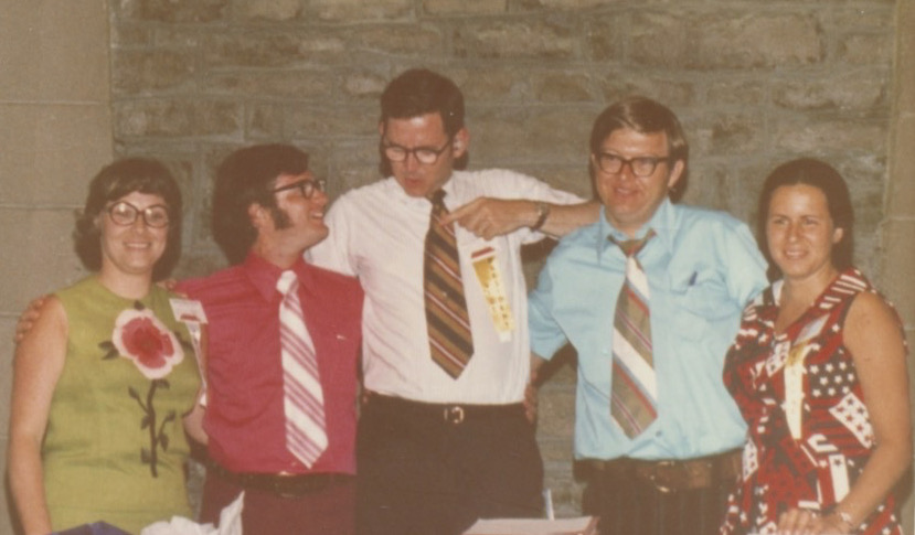 Five young white people stand with their arms around each other. The first woman has on a sleeveless green top with a large red flower in the middle of the shirt. Next to her is a young man in a red shirt and striped tie. Next to him is a taller young man in a white button-up shirt with a dark striped tie. To the right is a another, slightly shorter, white man in a blue shirt with a striped tie, and finally there is a young woman with her hair in braids wearing a sleeveless red dress with a patchwork pattern.