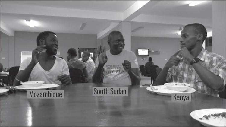 Three young black men in casual clothes are sitting at a table in a canteen with empty bowls and places in front of them. The man on the right, captioned Kenya, is signing with his right index finger on the middle knuckle of his closed left fist. The man in the middle, captioned South Sudan, is smiling at the man on the right, and has his right hand raised palm out, fingers loosely extended, thumb folded in, and his left hand in a closed fist in front of his chest. The man on the left, captioned Mozambique, is also smiling and watching the man on the right; his right hand is raised to beside his chin with the fingers loosely pinched.