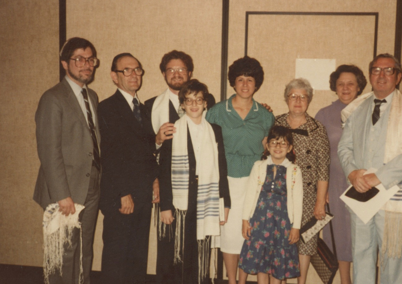 A group of nine white people standing in a room. The man on the far left is wearing a yarmulke and is holding a fringed white shawl. A teenage boy in the center is wearing a white and blue fringed shawl while his father, his hand on the boy's shoulder, also wears a blue and white shawl. The boy's mother stands next to the father in a green shirt. In front of her is a little girl in a blue and red dress with a white cardigan.