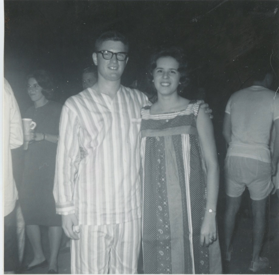 A black-and-white photo of two young white people. The man is dressed in vertically striped pajamas. He has large black glasses and a short hair cut. His arm is around a young woman with wavy chin-length hair, wearing a sleeveless vertically striped housecoat. 