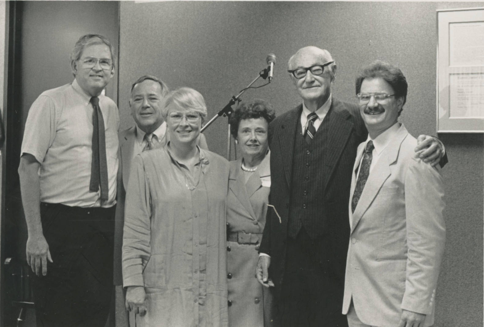A black-and-white photo of six white people. The first man on the left is very tall and wears a short sleeved button-up shirt with a skinny dark-colored tie. Next to him is a much shorter white man with gray hair. In front of him is a white woman with light hair in a pixie cut. Behind her is a shorter middle-aged woman with dark curly hair wearing a pearl necklace. Next to her is a tall, older white man with black-rimmed glasses in a dark suit. He has his arm around a younger white man with glasses and a mustache who's wearing a light colored suit. Behind the group is a microphone on a stand.