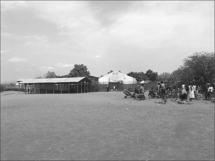 Exterior shot of the Food Distribution Center. The top of a tall canvas tent can be seen beyond a high wire fence. Inside the fence are some trees; outside is dry dirt, with a few trees in one corner and a wall-less wooden structure with a pitched corrugated iron roof beside the fence. Around 20 black people are waiting in near the trees on the right hand side of the picture, some standing, some sitting, some bending over bundles of sticks. Several have bicycles; there is also a motorbike and a wheelbarrow seemingly filled with bundles of sticks.