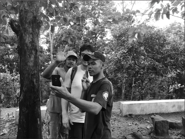 Three young Balinese men, two in baseball caps, one without a hat, smiling. One is waving to the camera. One man is holding a phone up as though photographing something to the side.