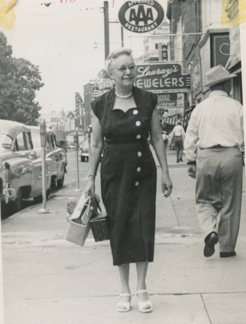 A black-and-white photo of an older white woman walking down a city sidewalk. She's wearing a dark colored dress with a sweetheart neckline large white buttons down the front. She's carrying two straw bags. Behind her are signs for AAA Restaurant and Lauray's Jewelers.