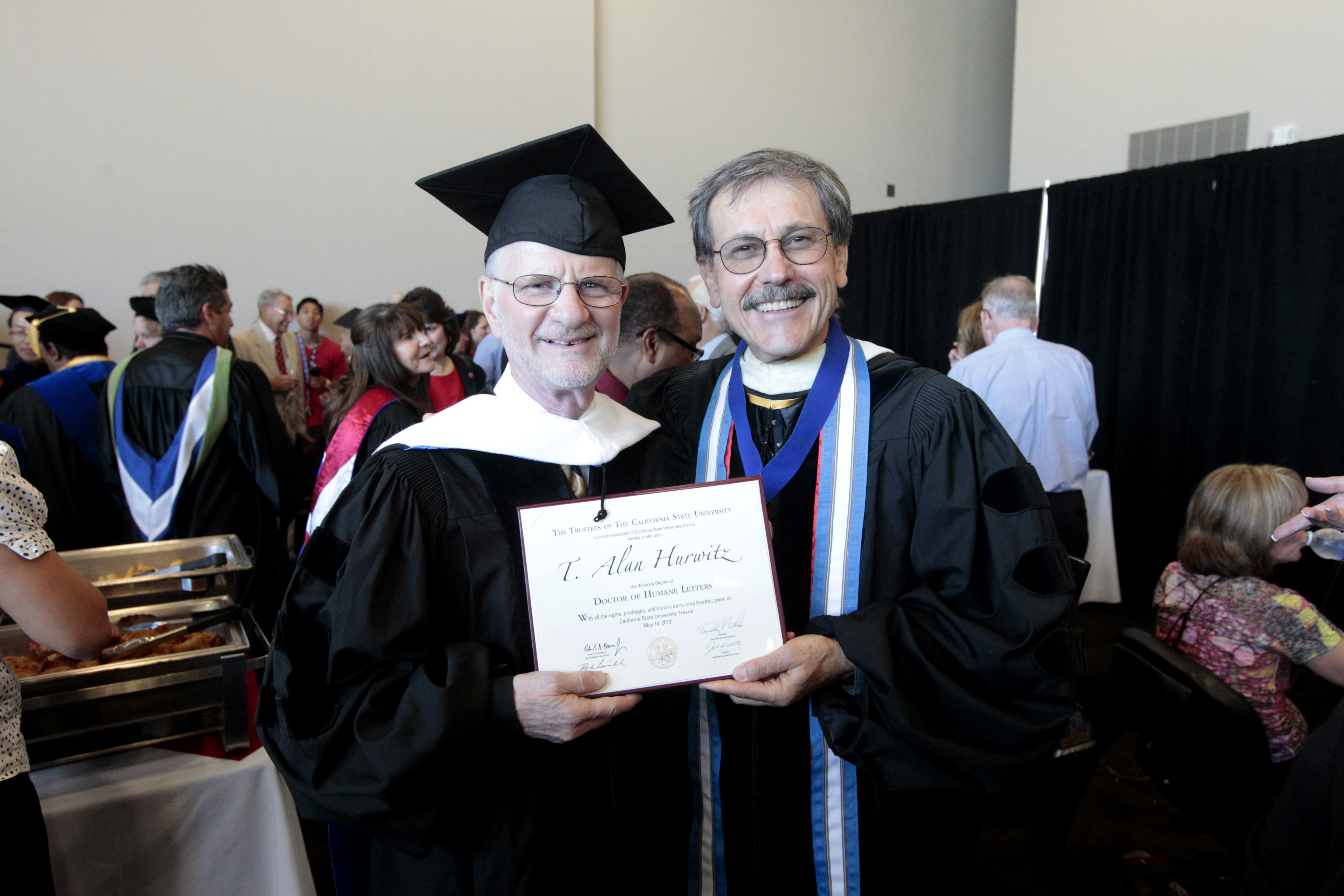 Two older white men both in dark robes. One is wearing a mortar board white the other is not but is wearing a light blue and white sash around his neck. They are both holding up a framed piece of paper with the words T. Alan Hurwitz written in calligraphic script across it.
