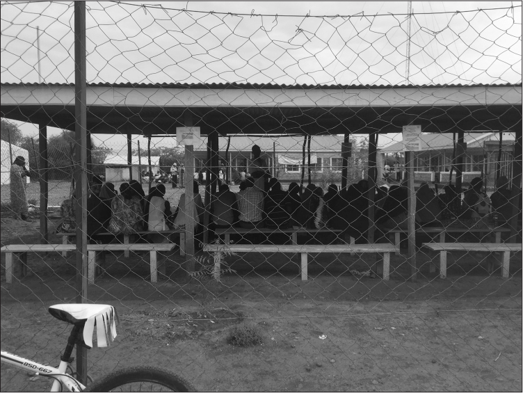 View through a chicken wire fence with a bike propped up against it. Inside is a wall-less metal or concrete structure with a flat corrugated iron roof. Inside are wooden benches with a large number of black people sitting on them.