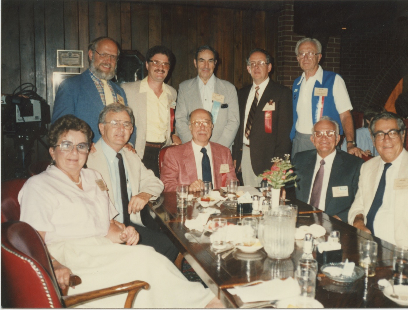 A group of middle aged and older white people (one woman and nine men) sit and stand around a dark wooden table full of glasses and used plates. All of the men save one are wearing name tags with ribbons. The woman is also wearing a beribboned name tag.