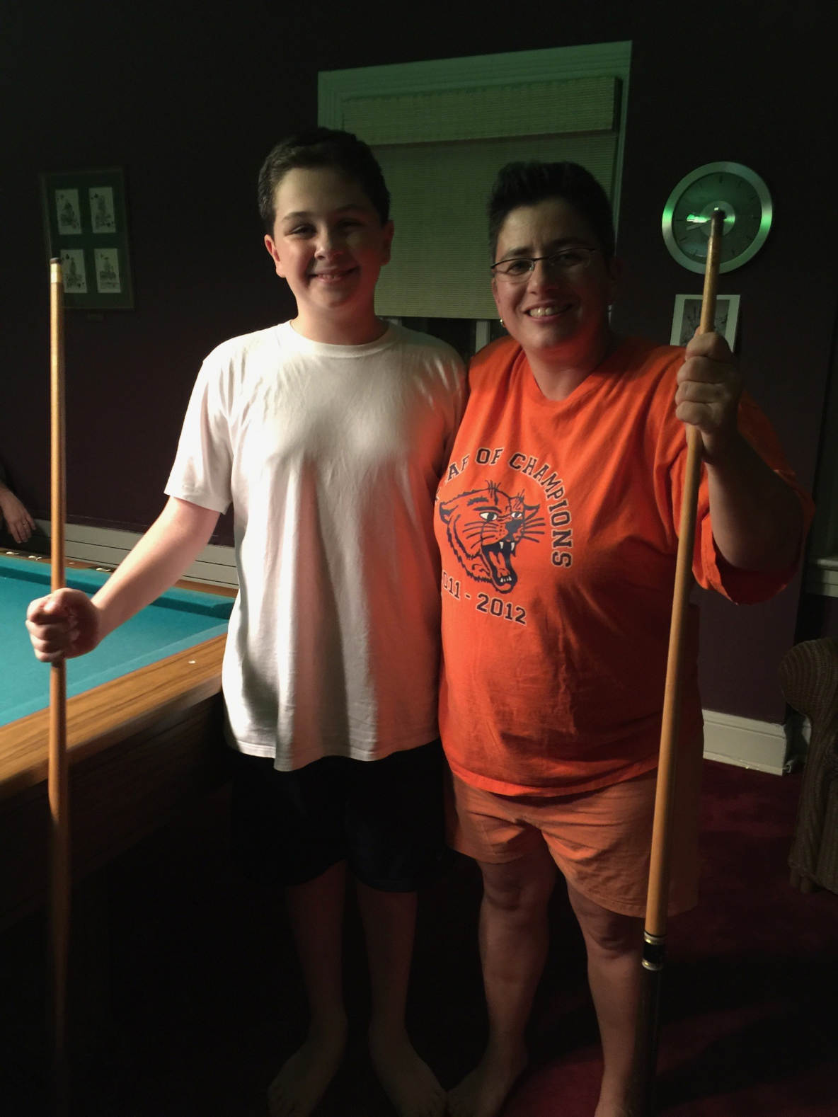 A young white teenage boy and a middle-aged white woman both holding pool cues, stand with their arms around each other next to a pool table.
