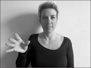 Annelies, a white woman with short blonde hair, signs COUNTRY (right hand in front of right shoulder, hand open and clawed, palm facing the camera).