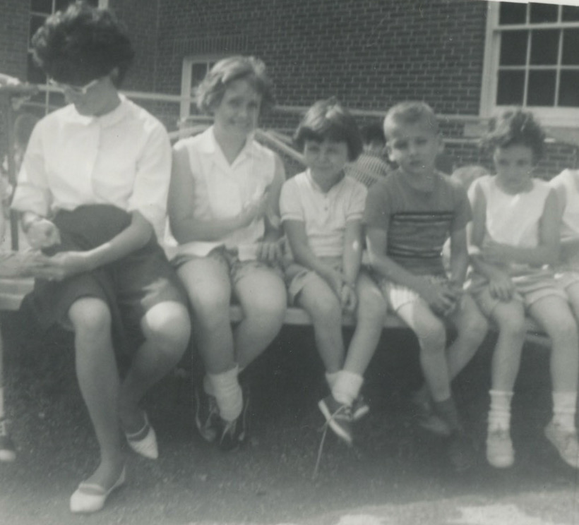 A young white woman sits on a bench outside of a brick building. Next to her are two white girls with bobbed hair. Next to there is a tow-headed white boy. Next to him is another little white girl. The young woman is turned away from the camera and is looking down like she might be talking to another child just out of the frame.