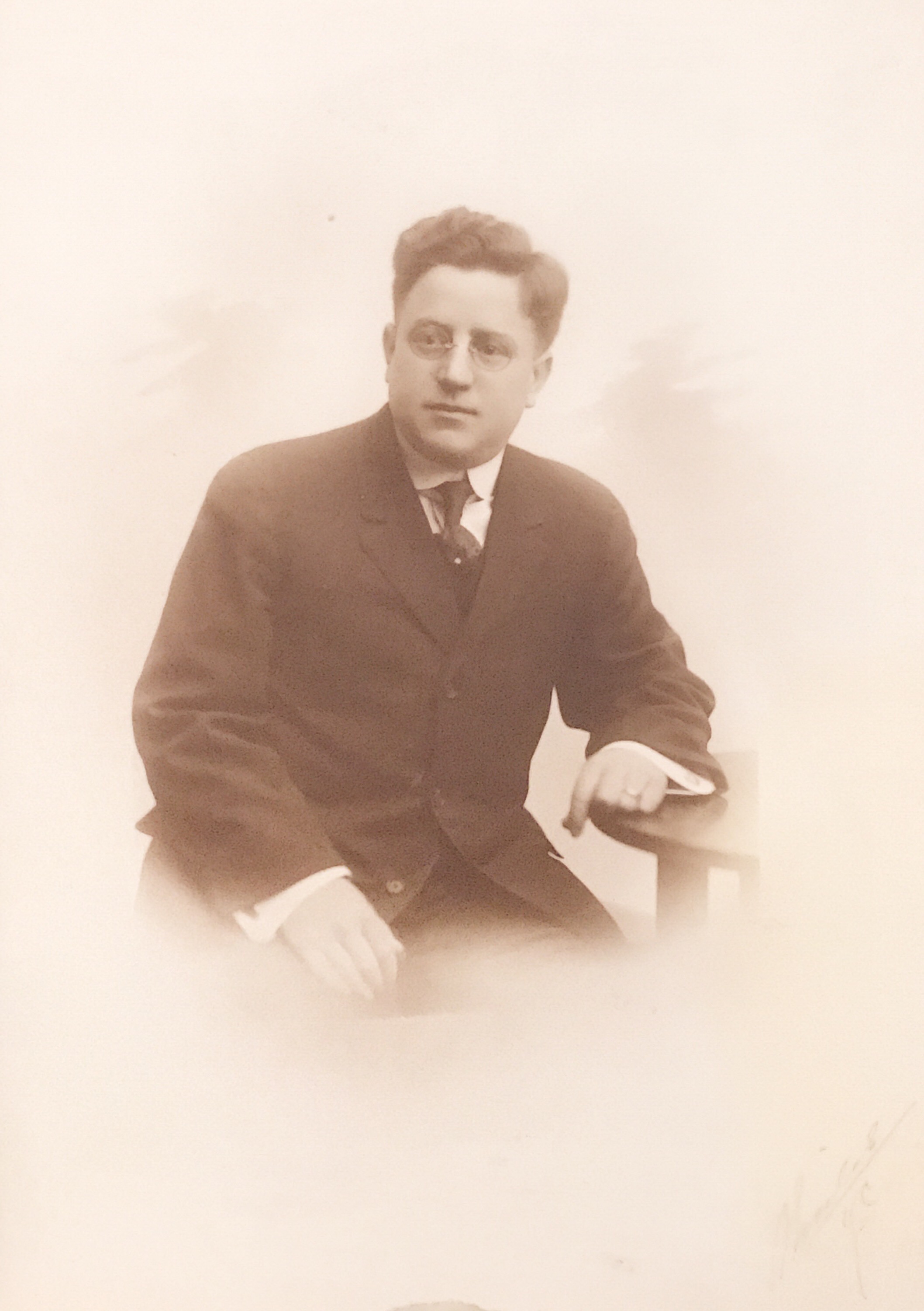 A sepia-colored photo of a young white man in glasses and in a dark-colored suit with a dark-colored tie. His dark hair is wavy and cut short. He's sitting down with one arm leaning against a table.