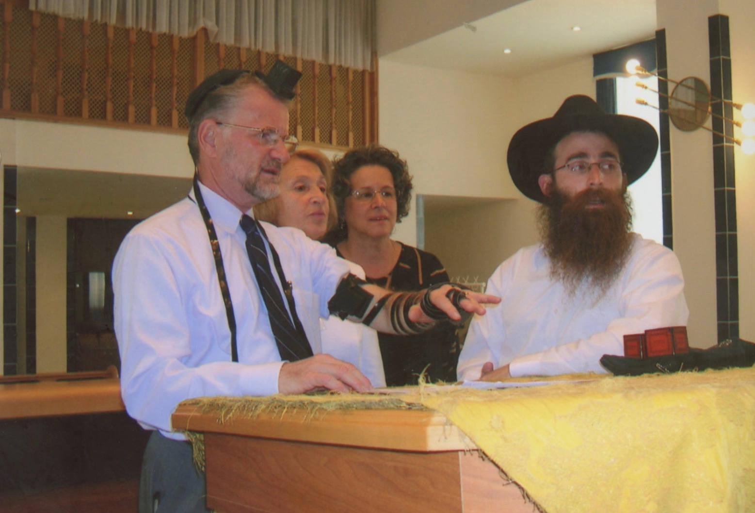 A white man in a white shirt wearing Tefillin (a cubic black leather boxes with leather straps) on his head stands at a podium next to a rabbi with a black wide brimmed hat and a bushy beard. Behind them are an older woman with reddish hair and a middle-aged woman with dark wavy hair. The man wearing Tefillin is extending his arm out white the rabbi leans one elbow on the podium.