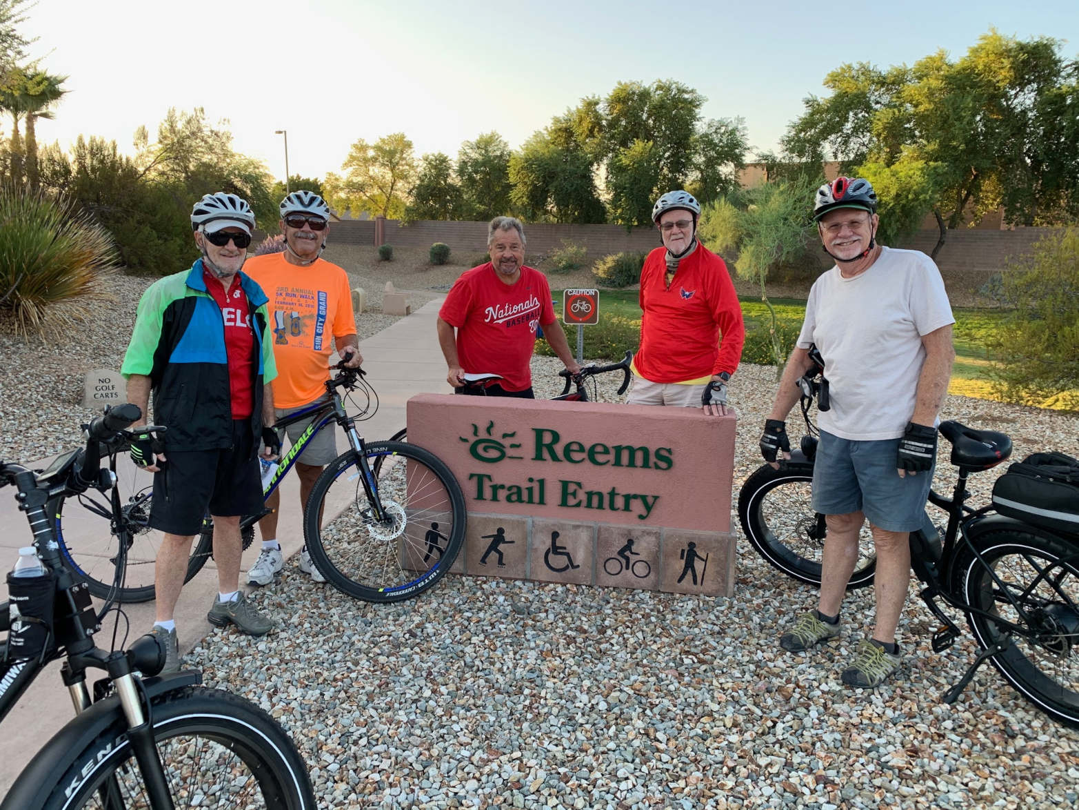 A group of older white bicyclists at a trail head. They are standing with their bikes in front of a sign that reads Reems Trail Entry.