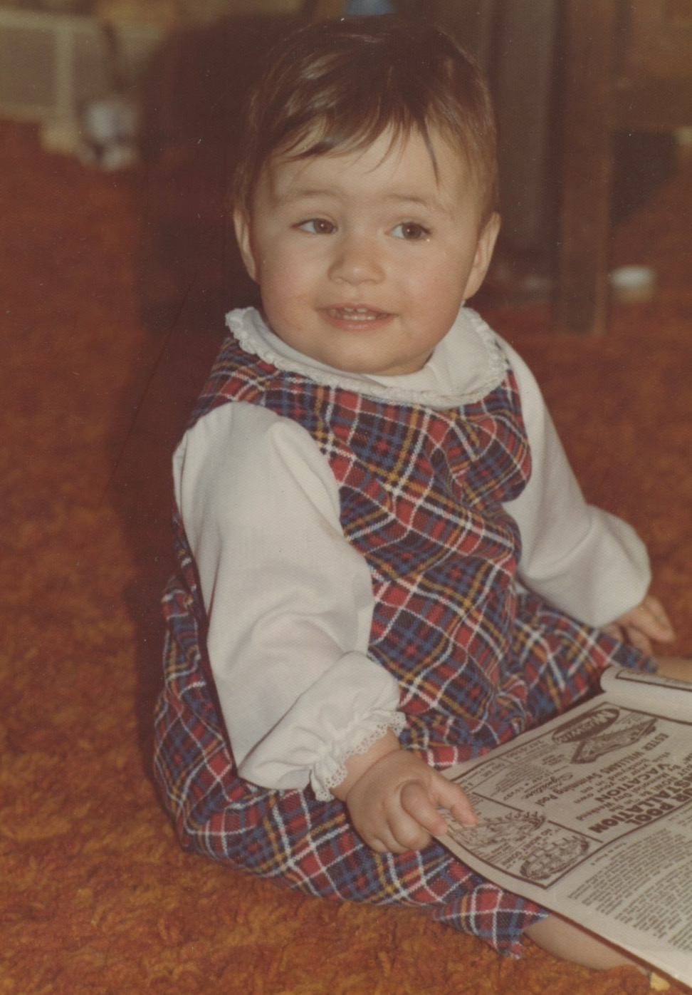 A baby white girl with rosy cheeks sits on the floor in a red, blue, white, and yellow plaid jumper with a white blouse underneath. she has short brown hair and is smiling.