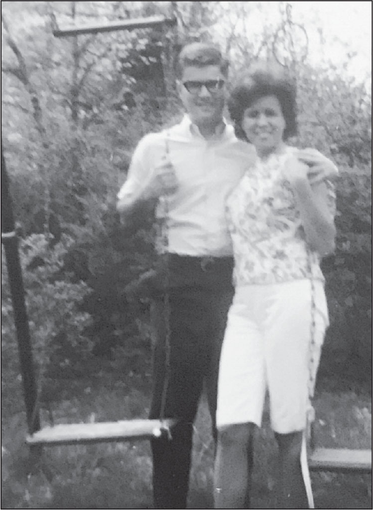 A young white man and woman, smiling at the camera, stand side-by-side with their arms around each other. They both are holding onto chains of playground swings.