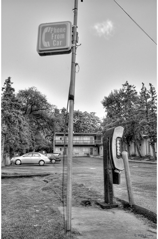 black-and-white vertical photograph of a sign high up on an electric pole. The sign reads: “Phone From Car.” Next to the sign is a drive-up phone booth in the shape of a telephone receiver. In the background are some cars parked at a motel.