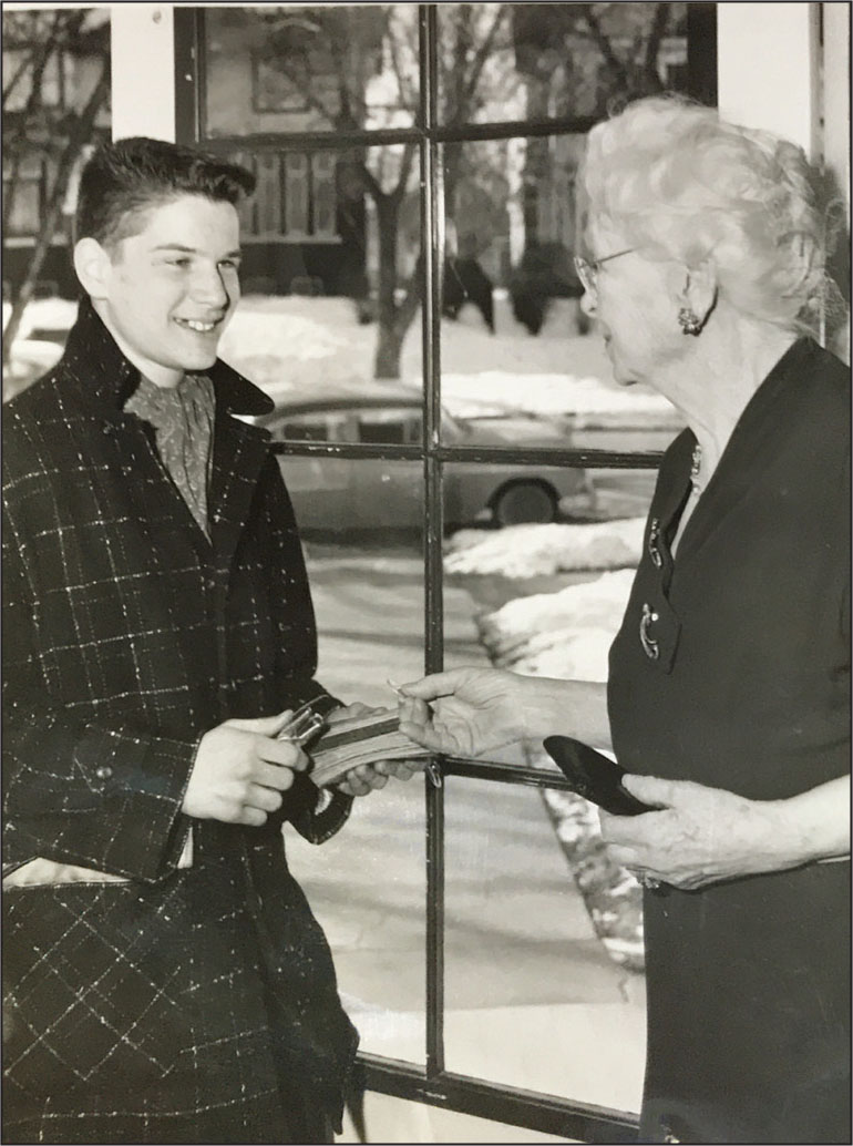 A young white teenage boy wearing a thick, checkered coat and a scarf stands in front of the front door of a woman's house. The elderly, white-haired woman holds a wallet and appears to be giving the boy some money. He holds a one-hole paper punch. Behind him a car is parked on a snowy street.