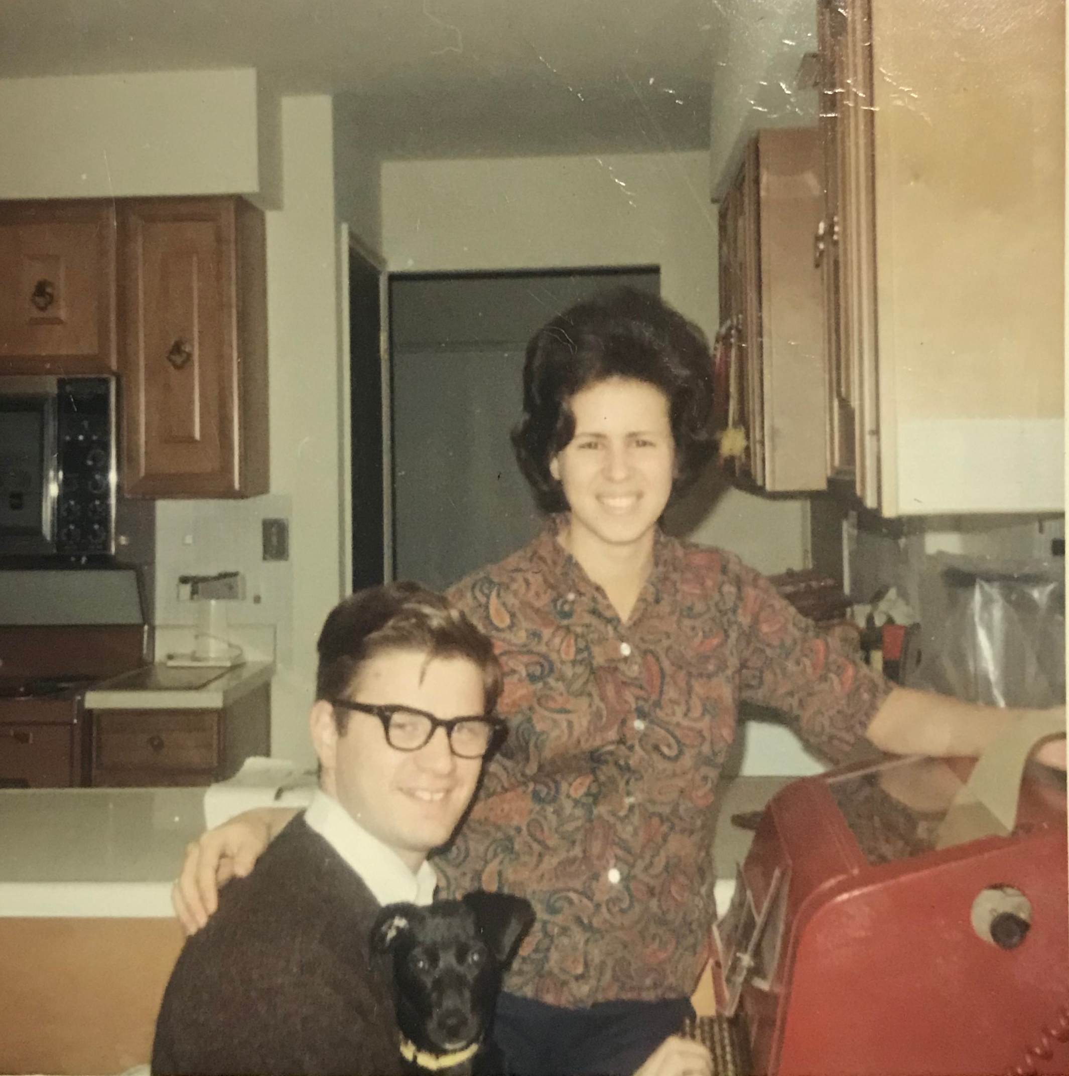 A young white man with horn-rimmed glasses sits in front of a red teletypewriter with a black dog in his lap. A young white woman with a dark bouffant harido stands next to him with her hand on his back. Behind them are kitchen cabinets and a counter.