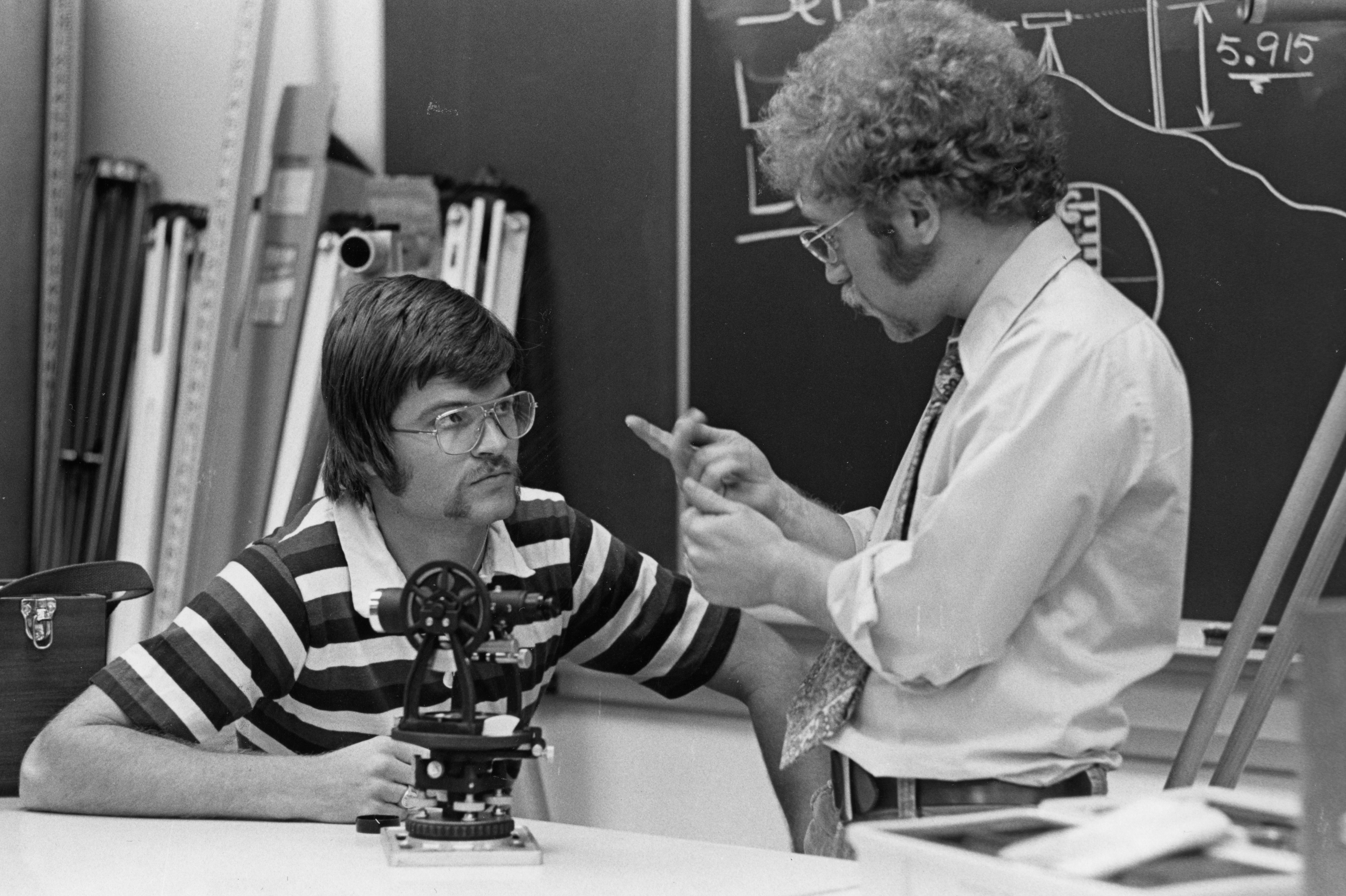Black-and-white photo of a young man with glasses sitting in front of a blackboard and looking at an older man with curly hair and side burns explain something to him in sign language.