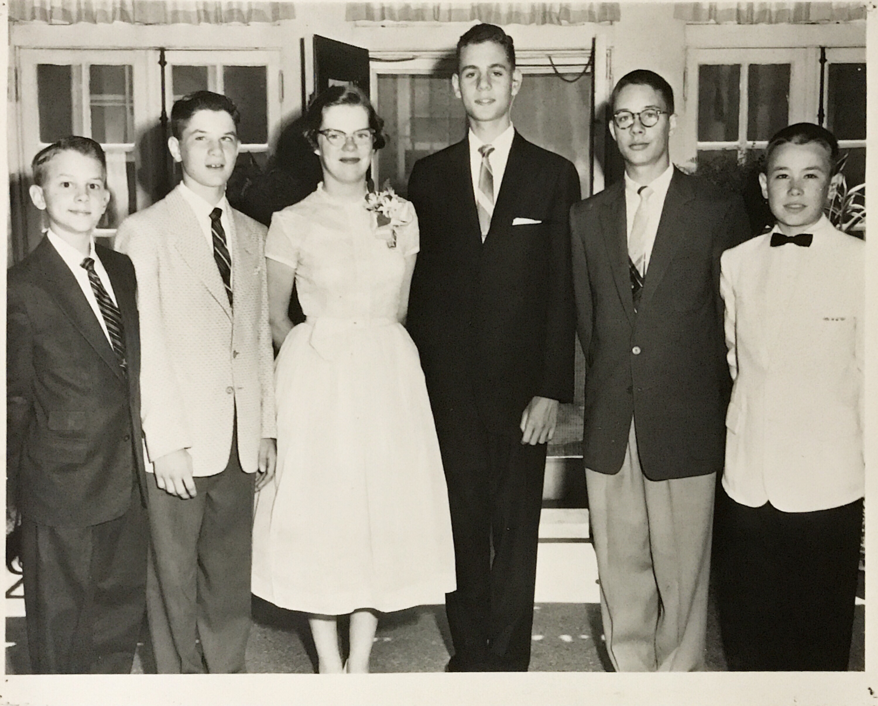 A black-and-white photo of six young white people--five boys and one girl. They are all dressed up--the boys in suites and ties and the girl in a lgiht-colored dress with a cinched waist. The girl is wearin a corsage pinned to her dress's neckline.