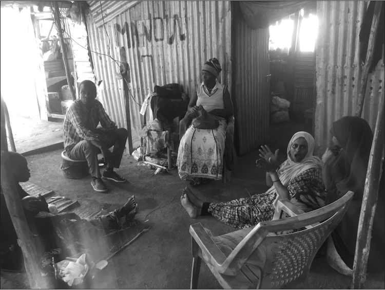 Interior of a corrugated iron hut. Six people (including women wearing chadors/headscarves, one man in a checked shirt and trousers, and a baby) sit on plastic chairs, buckets, and the dirt floor. One woman is looking at the camera and signing.