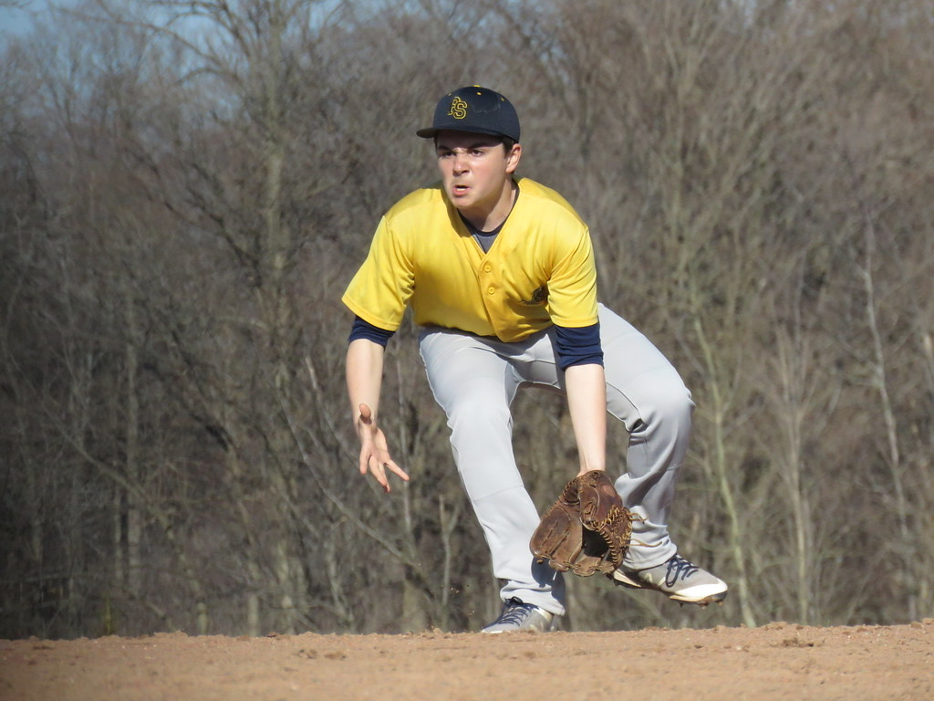 A young white man in a yellow and gray baseball uniform with a determined and focused expression crouches down to catch a ball in his baseball mitt.
