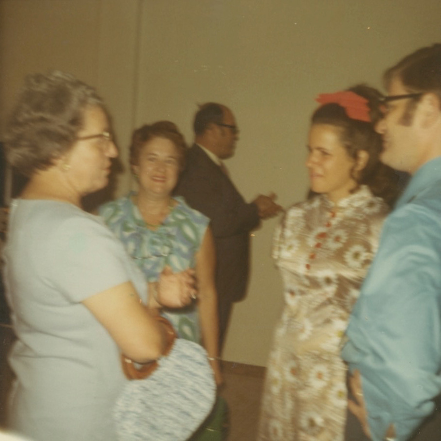Blurry photo of a older white woman with glasses in a blue dress talking to an older white woman in a flowerd dress and a young white woman with a red bow in her hair. The younger woman is wearing Cheongsam style dress with a high collar and red buttons down the front. A young white man in a blue shirt and glasses stands next to her.