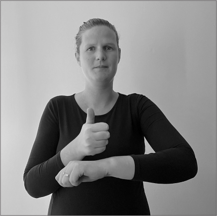 Amandine, a white woman with fair hair pulled back off her face, signs BLAME (left elbow bent at 90 degrees, left arm crossing her torso at stomach level, left hand in fist palm downwards; right arm bent at elbow, right fist above left hand, palm inwards, thumb raised).