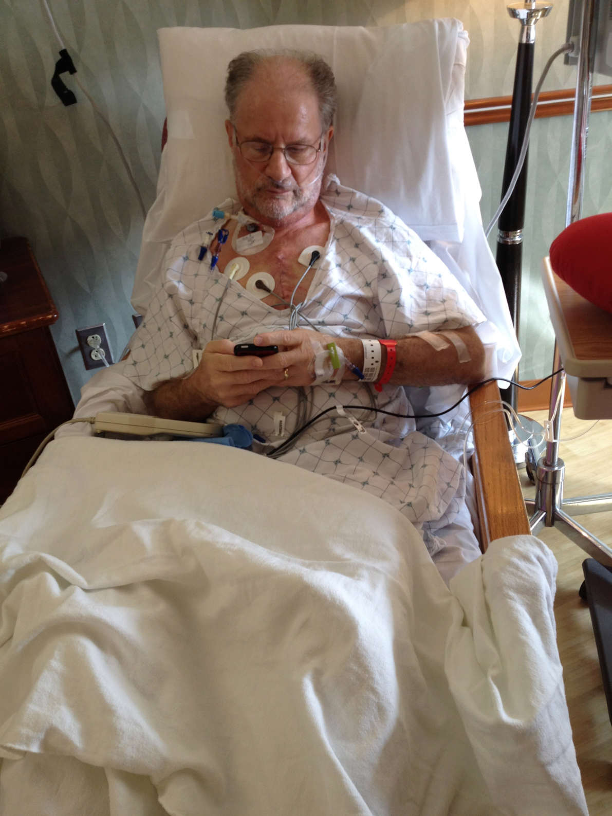 An older white man in a hospital gown sitting in a bed looking at his cell phone. He's has a fresh incision and electrodes attached to his chest. There is an IV going into the back of his hand.