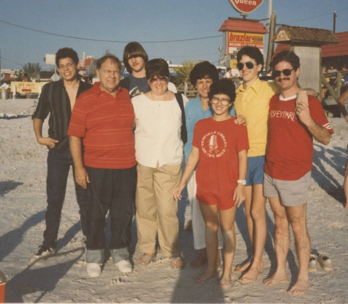 Two white families standing on a sandy beach. The children--three boys and one girl--are all teenagers. The family of four on the left are all wearing long pants and shoes while most of the other family of four on the right wear shorts and have bare feet.