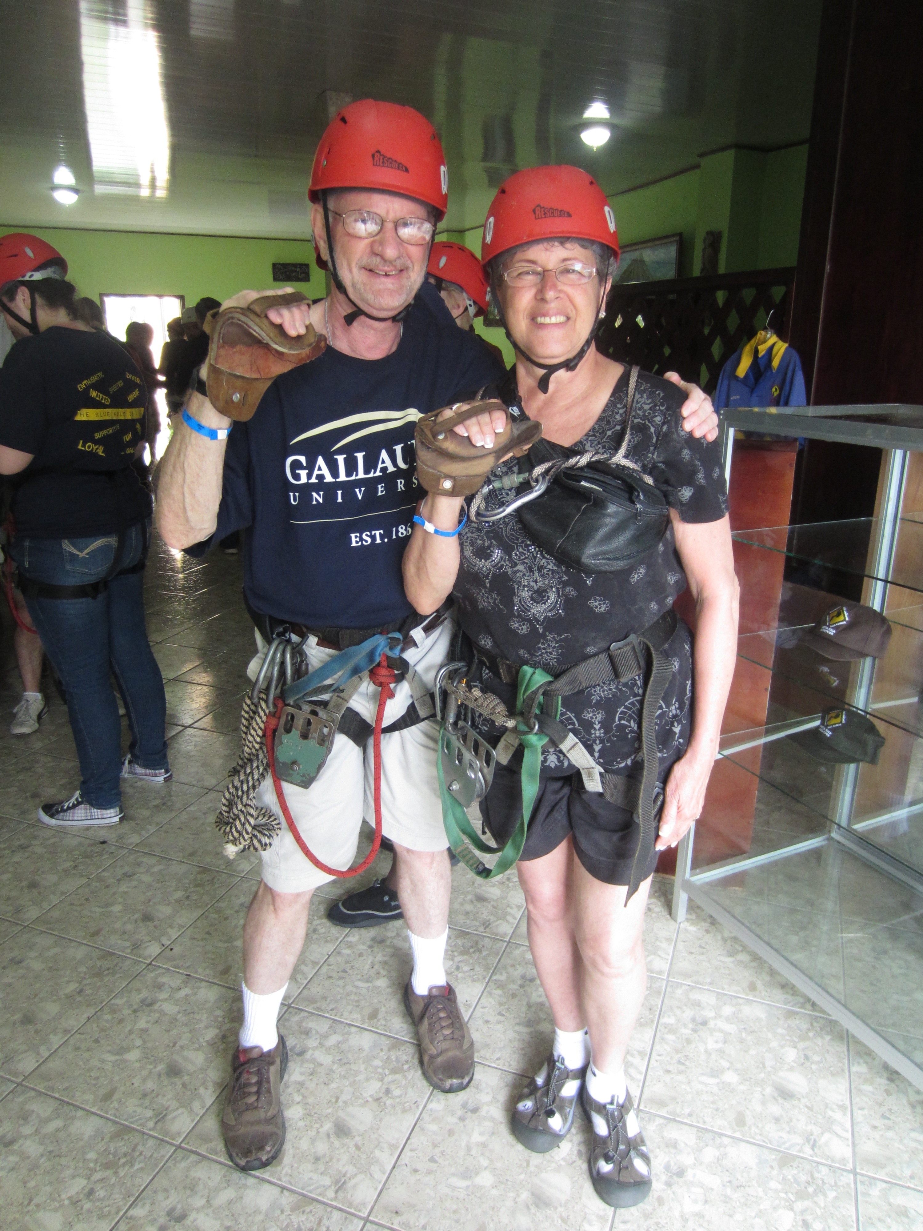 An older white man and woman both wearing harnesses, red helmets and thick leather pads on their hands. The man is wearing a dark blue t-shirt with the words Gallaudet University across the chest. The woman is wearing a black and white top. Behind them are other people in red helmets.