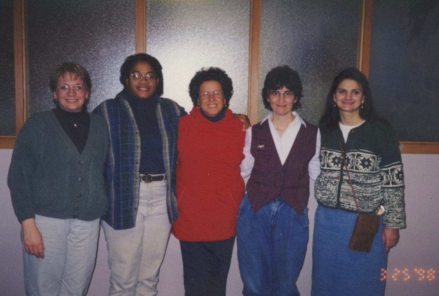 A group of five women standing with their arms around each other. The first woman on the left is a middle-aged white woman with glasses wearing a green cardigan over a black shirt. Next to her is a tall young Black woman also with glasses wearing a blue and gray unbuttoned shirt over a black shirt. To the right is a middle-age white woman with dark wavy hair, wearing glasses and a bright red top. Next to her is a white womn in a purple vest over a white shirt. Anf finally, there is a young white woman wearing a green Fair islae sweater with a cross-body purse. 