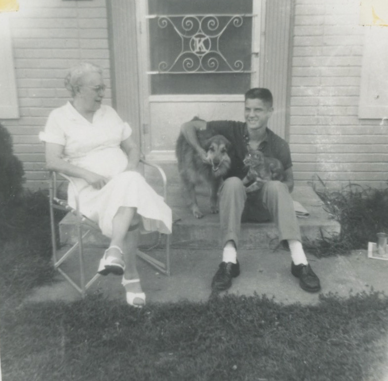 An elderly white woman sits on a lawnchair on a concrete walkway in front of a door with a scrolled screen featuring the letter K in the middle. A young white man with a dark crew cut sits on the stoop next to her holding a shepherd dog and a tabby cat. The woman is wearing a light colored short sleeved dress with white wedge heels.