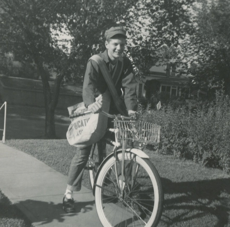 A white teenage boy stands astride a bike on a sidewalk with a metal basket affixed to its handlebars. He's wearing a cap, a jacket, and jeans, cuffed at the bottom. He has two large canvas bags strapped across both sides of his chest.