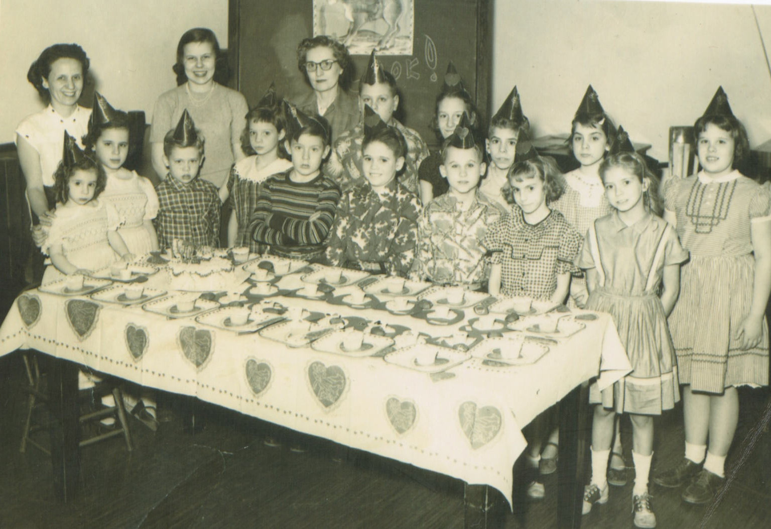 Three middle-aged white women stand behind a group of white children wearing party hats. The children are all standing behind a long table that has a table cloth festooned with hearts. On the table are cupcakes.
